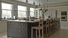 A large modern kitchen with tiled flooring, white cabinets, and a large kitchen island with grey drawers and a wooden countertop. Behind the island are seven tall wooden chairs facing large windows.