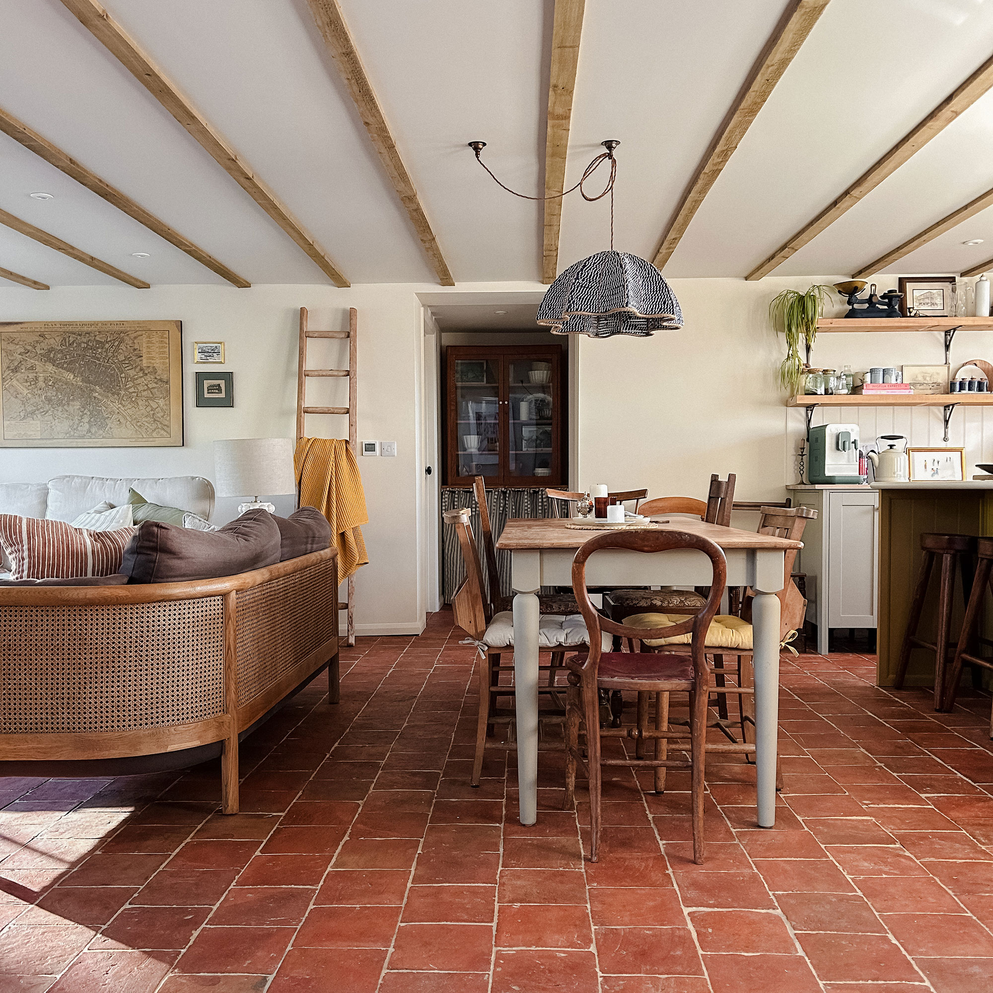 Open plan kitchen-dining-living space with terracotta tiled floor, slimline wooden ceiling beams, cabinetry, wooden dining table and chairs and cane-backed sofa