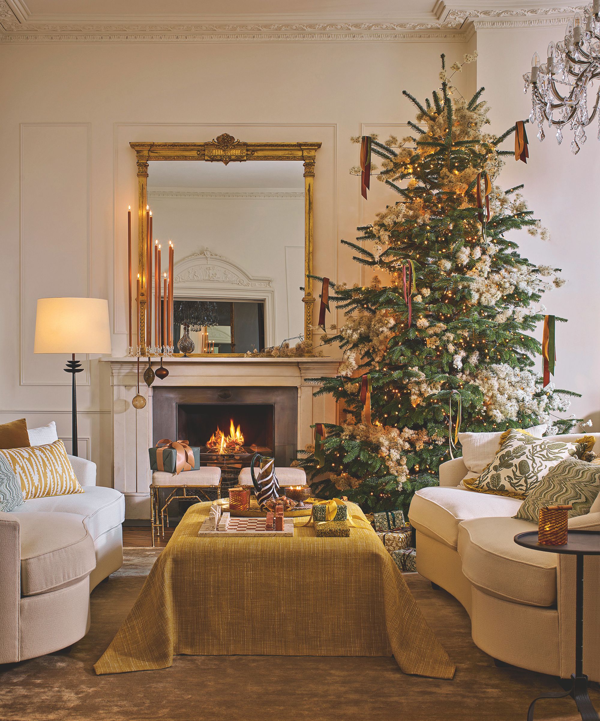 Luxurious Christmas living room with cream walls and furniture and an ottoman covered in a deep yellow throw. A large gold mirror is above the fireplace and a large tree decorated with golds and greens is in one corner