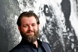 a bearded man in a suit smiles on a red carpet during a movie premiere event