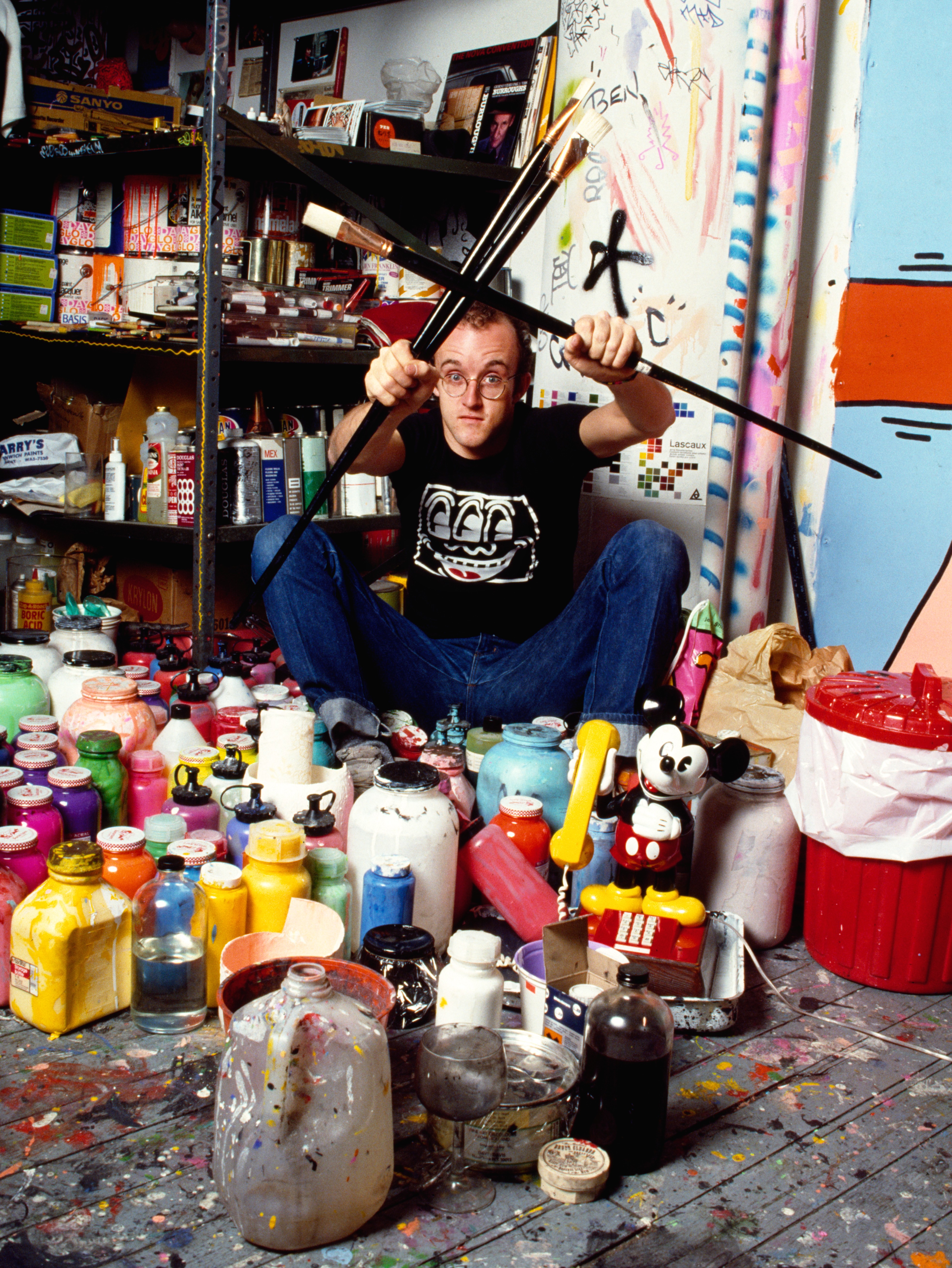 Colour photograph of artist Keith Haring seated among dozens of paint bottles and jars in a chaotic studio, holding multiple large paintbrushes crossed in front of him.