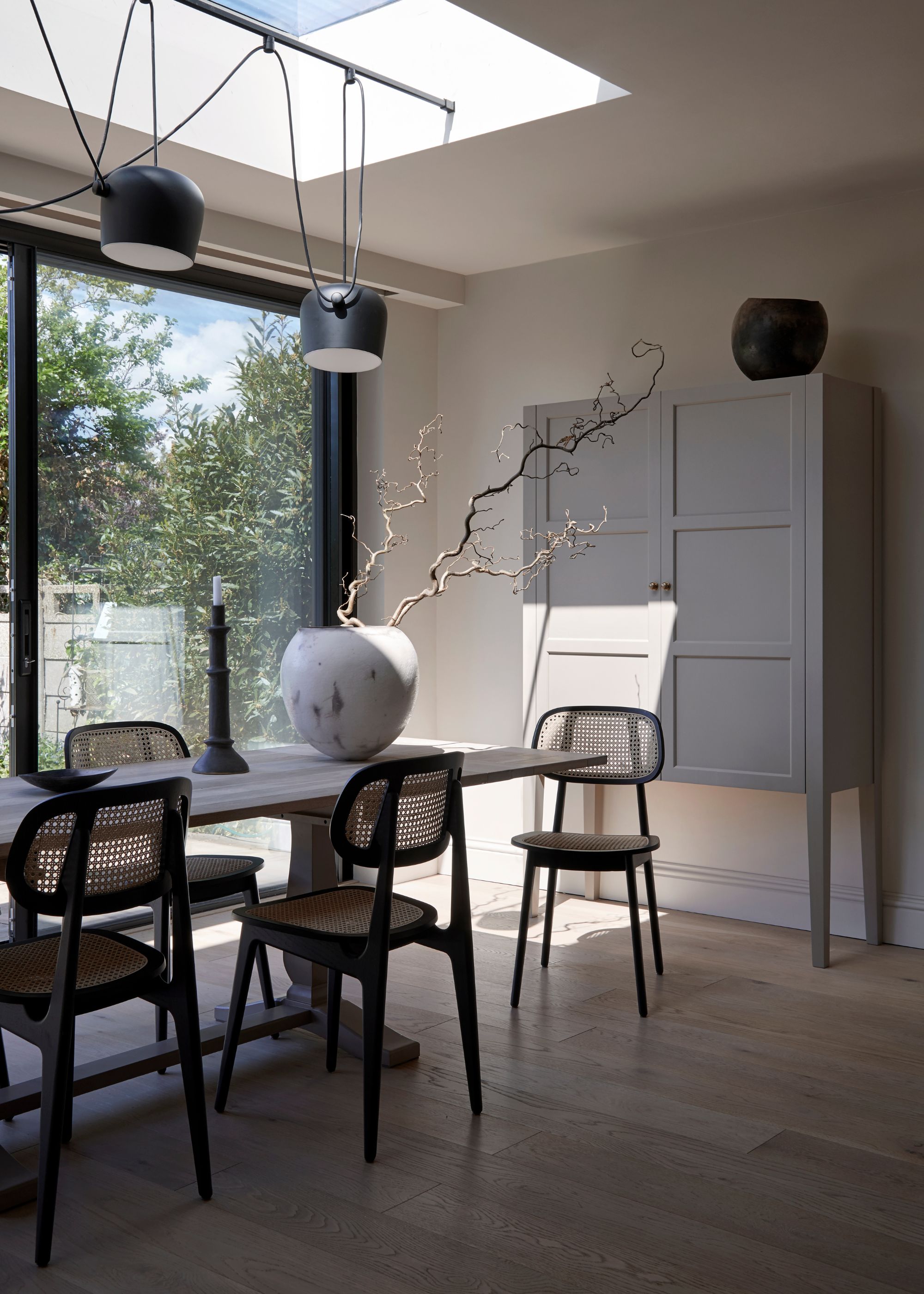 A kitchen with a dining table in the middle with chairs placed all around it and a large vase in the middle of the table. Above the are pendant lights with hanging cables.
