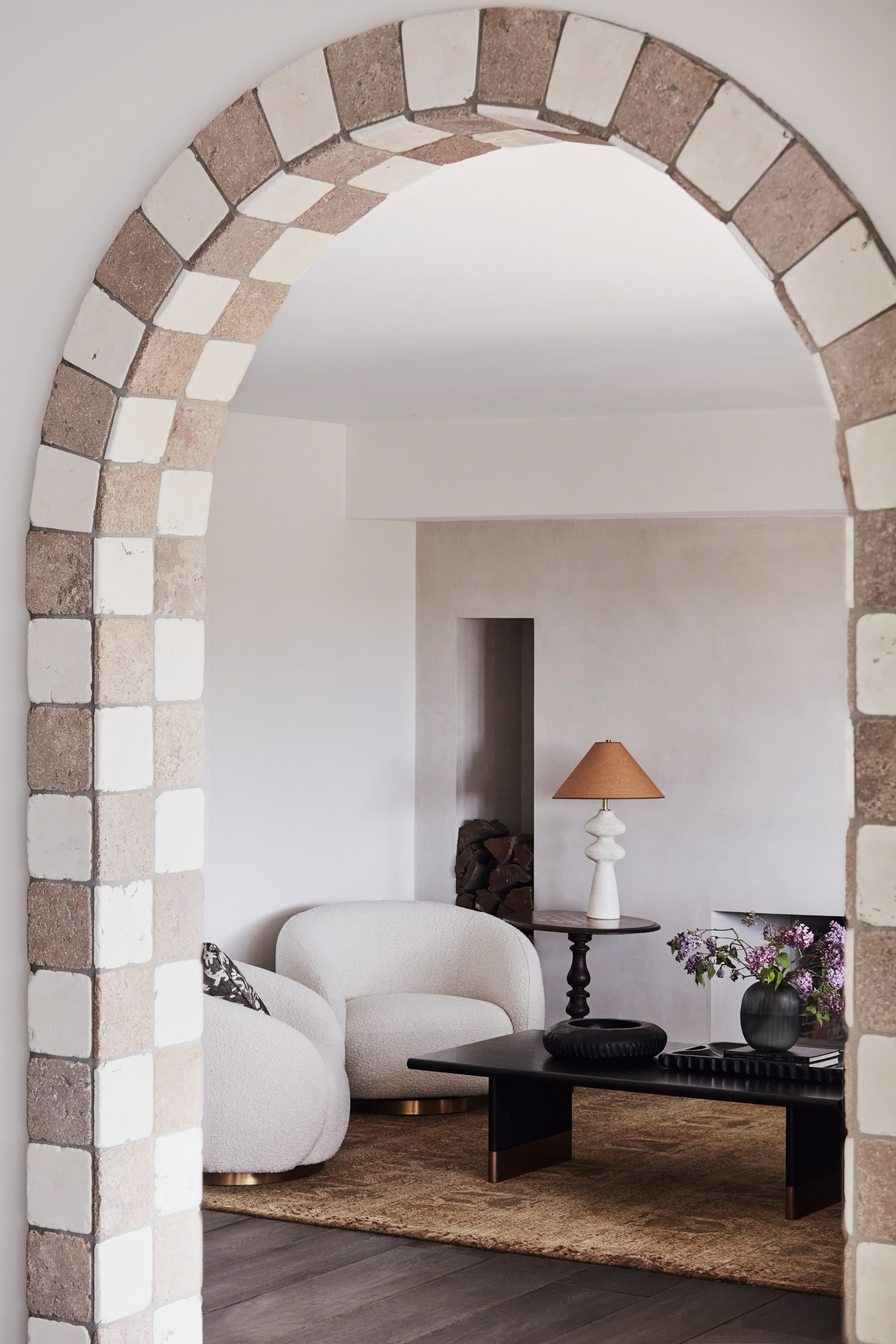 Arched doorway framed by checkerboard travertine tiles, leading to white living room with white curved armchairs and black coffee table