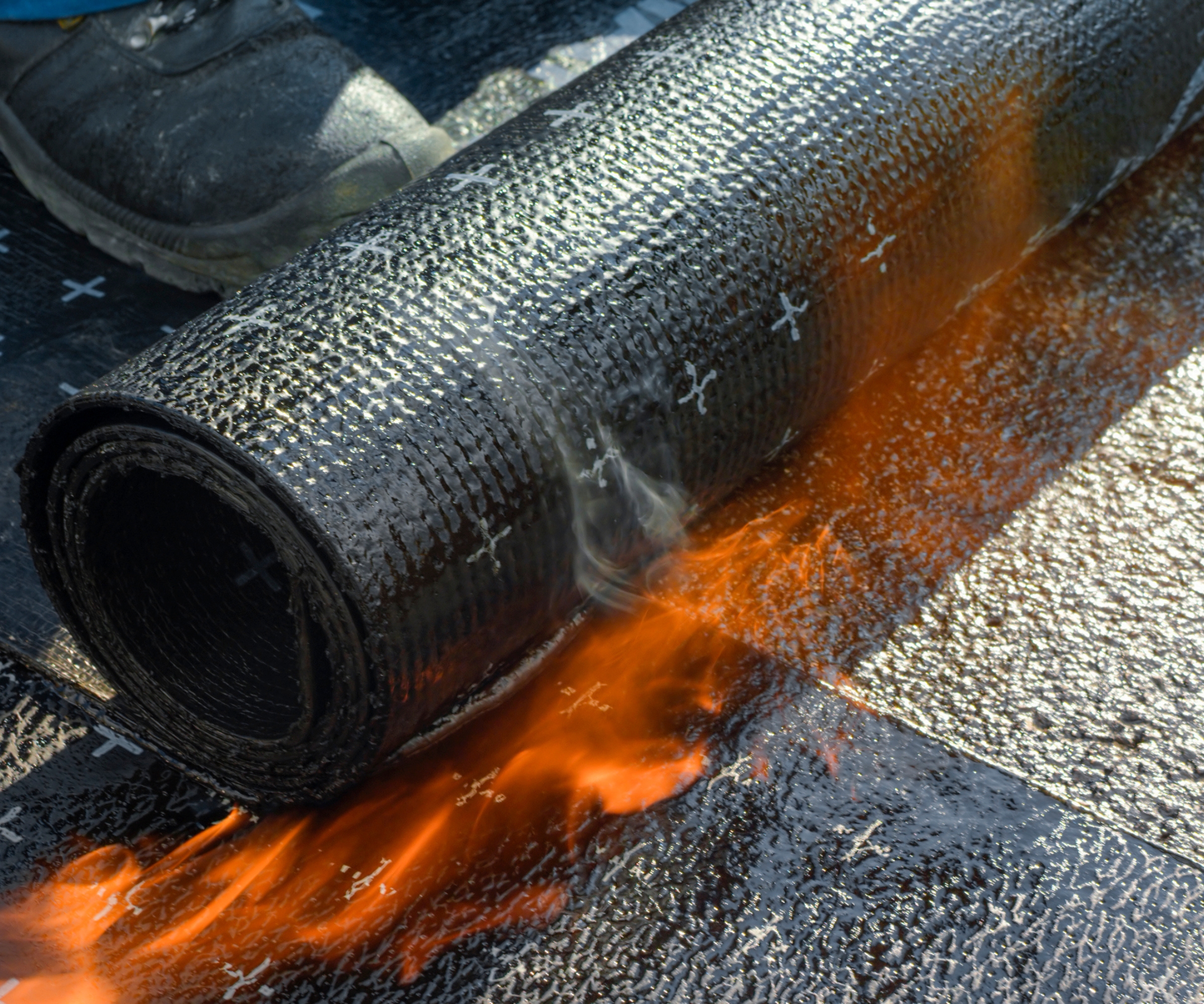 Rolled bitumen roofing being heated, while a roofer carefully rolls it out.
