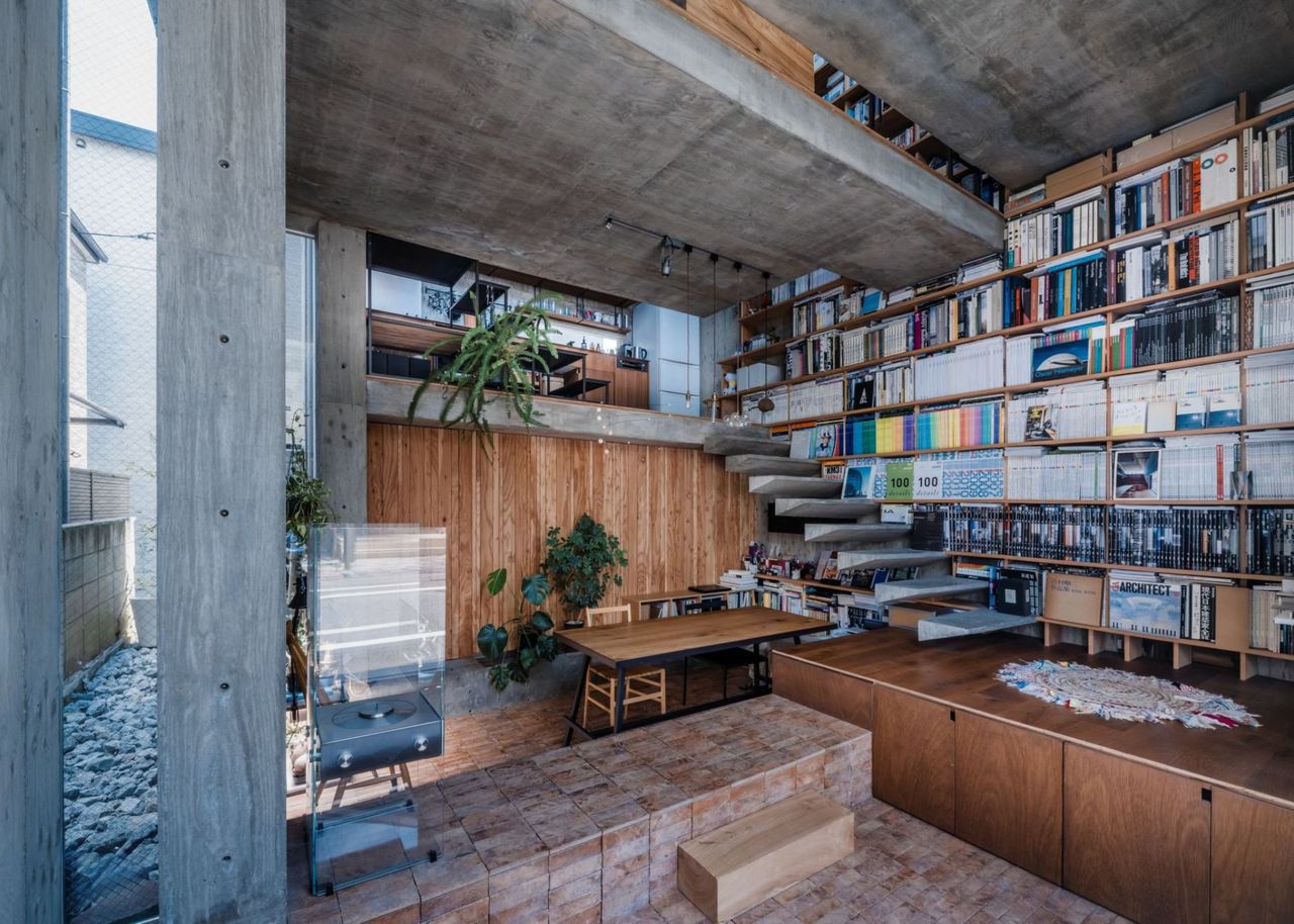 An open multi-layered Japanese home with concrete walls and ceiling, a bookcase that takes up the entire wall on one side, three stories up, with floating stairs and rooms with no doors