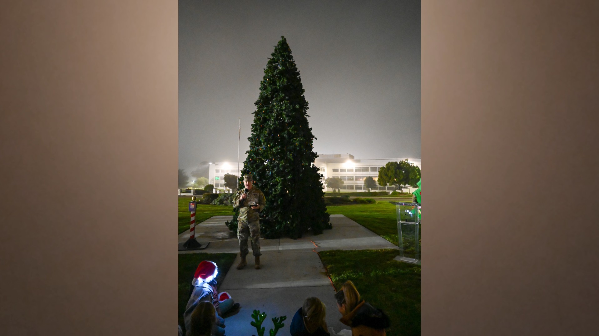 un hombre con uniforme de camuflaje habla por un micrófono frente a un árbol de hoja perenne en forma de cono