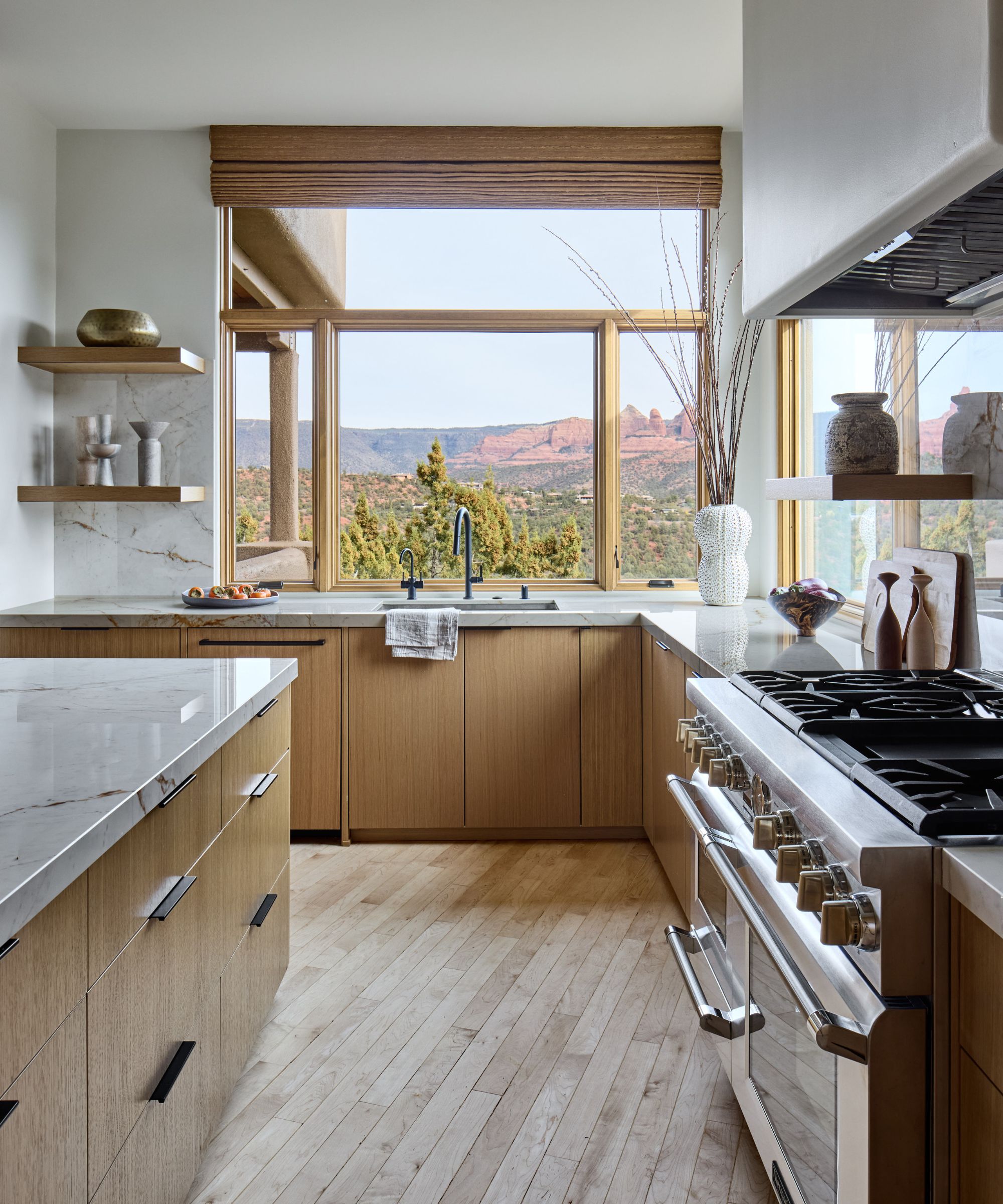 Small kitchen with wooden cabinetry and floor, wooden framed windows looking out to hills and greenery.