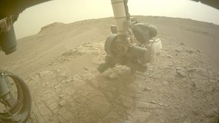 perseverance rover sampling head on the surface of mars. the view is very dusty and a large plane of rocks is in the front