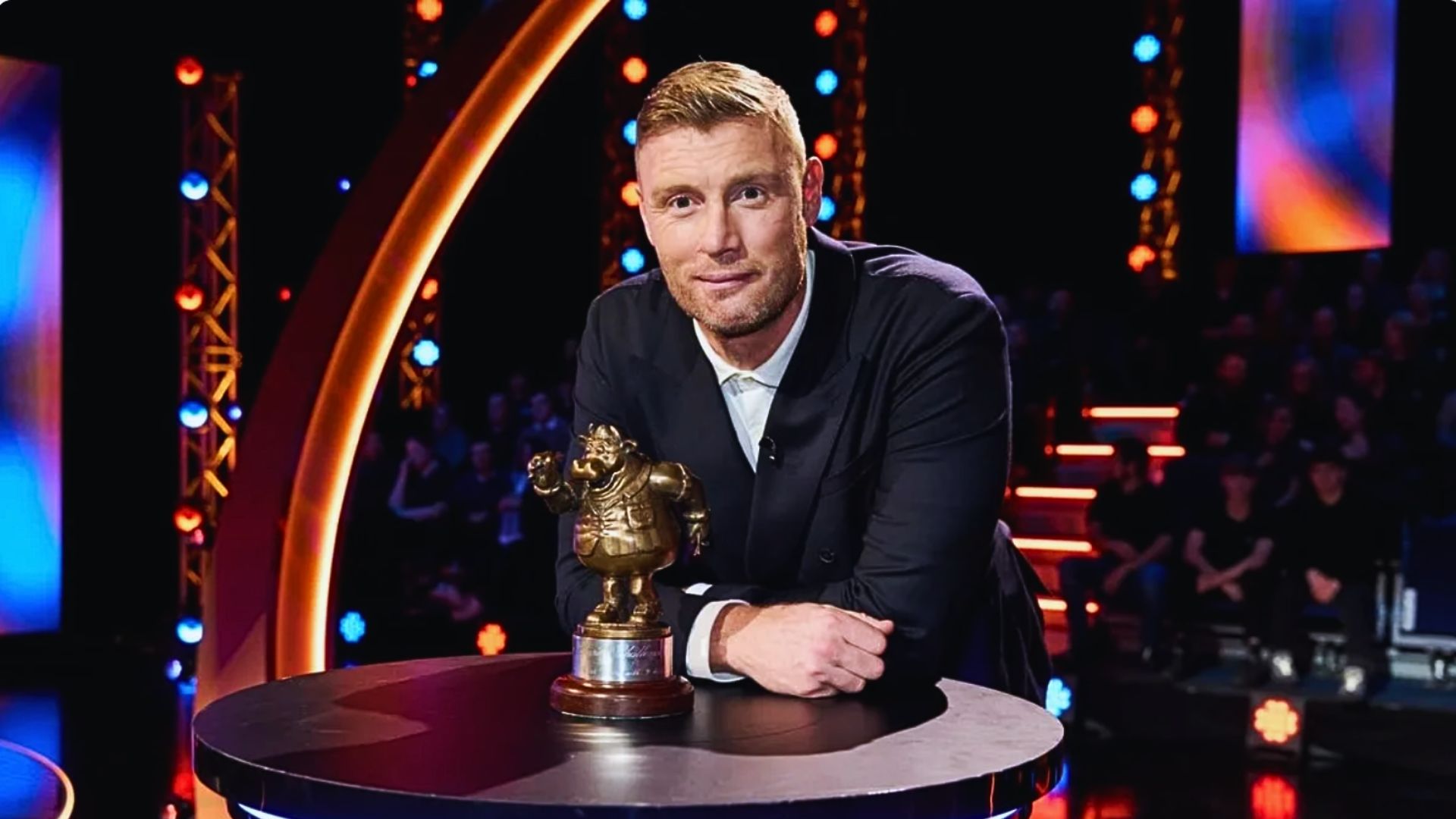 a promotional image of freddie flintoff for bullseye leaning on a table with the bullseye trophy in front of him