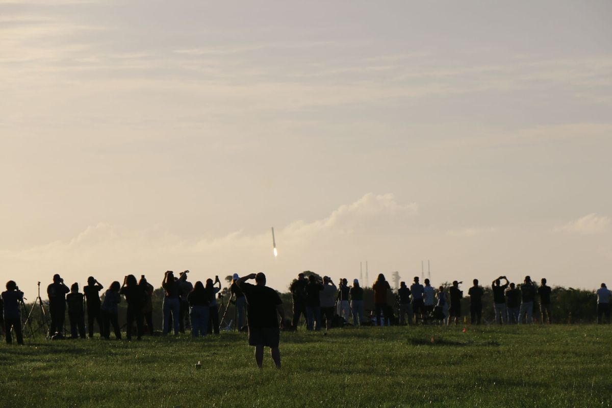 People stand on a lawn watching a long structure descend to Earth.