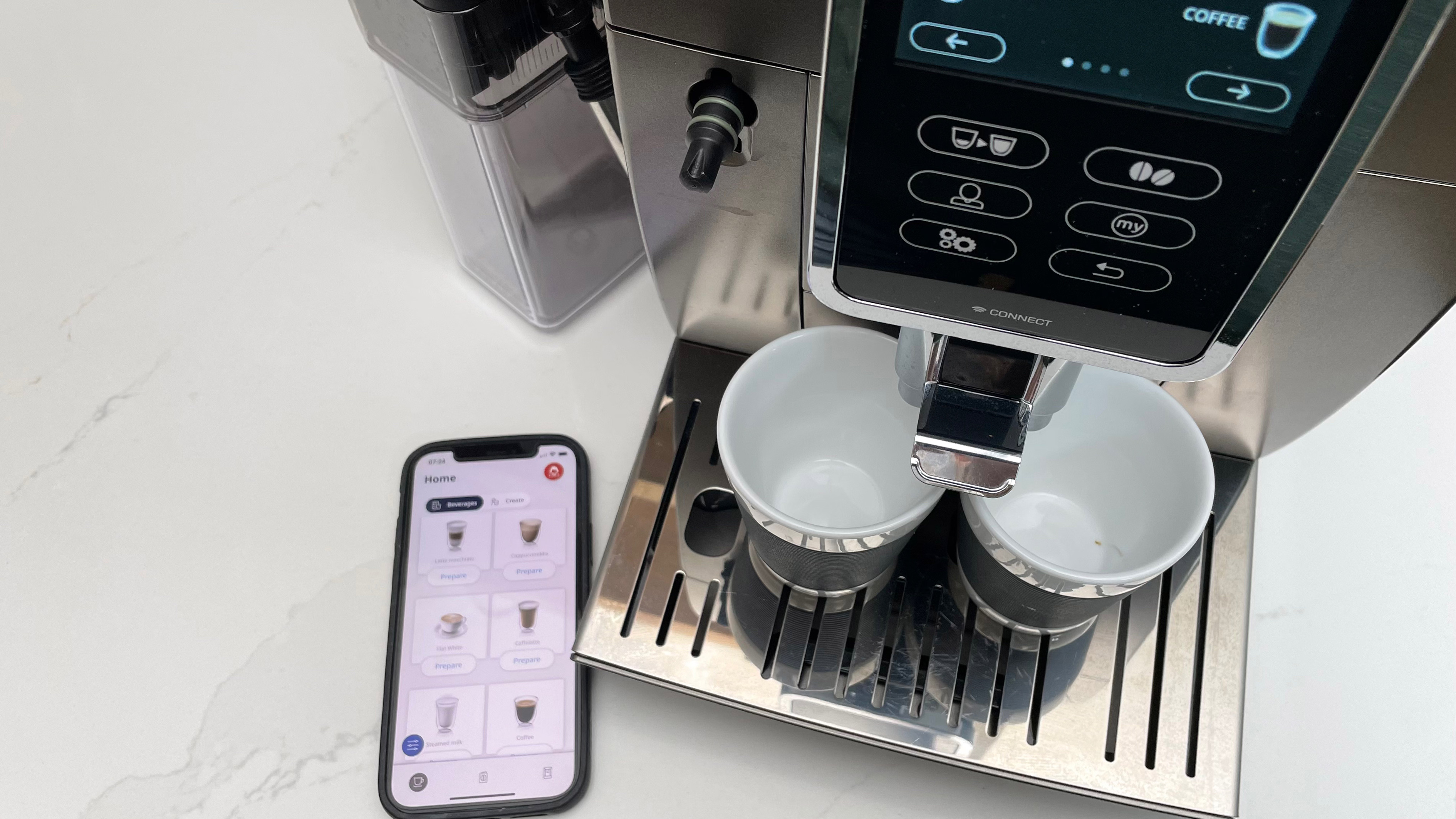 Looking down onto the De’Longhi Dinamica Plus as its ready to paur an espresso with the app onpen on a smartphone to the left-hand side