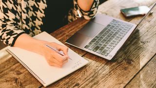 Woman writing in a journal next to laptop on desk