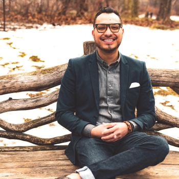 a man in a blue suit jacket, jeans, and black glasses sit cross legged and smiling at the camera