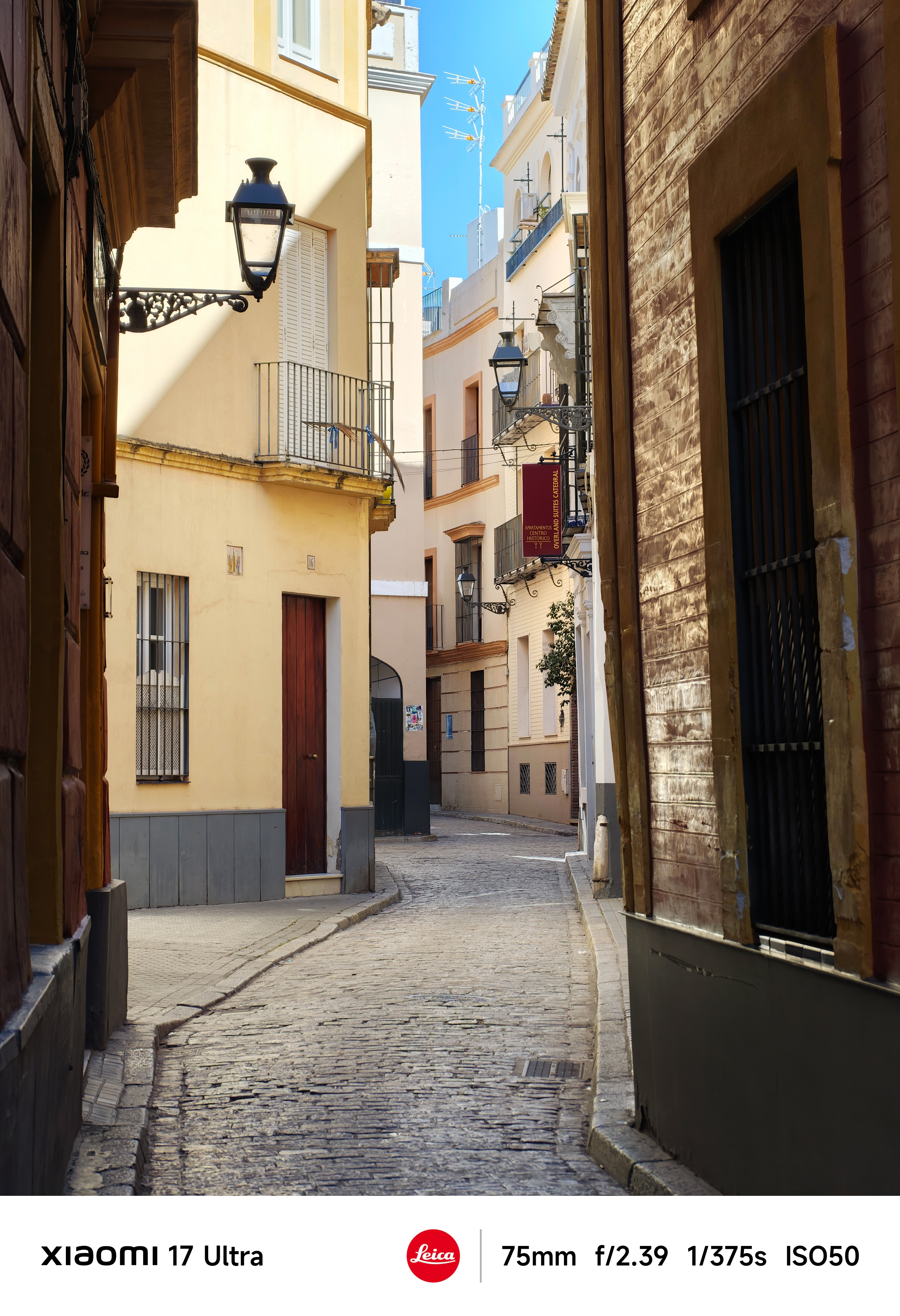 Narrow cobbled street lined with cream and ochre buildings, wrought-iron balconies and wall-mounted lanterns under a clear blue sky.