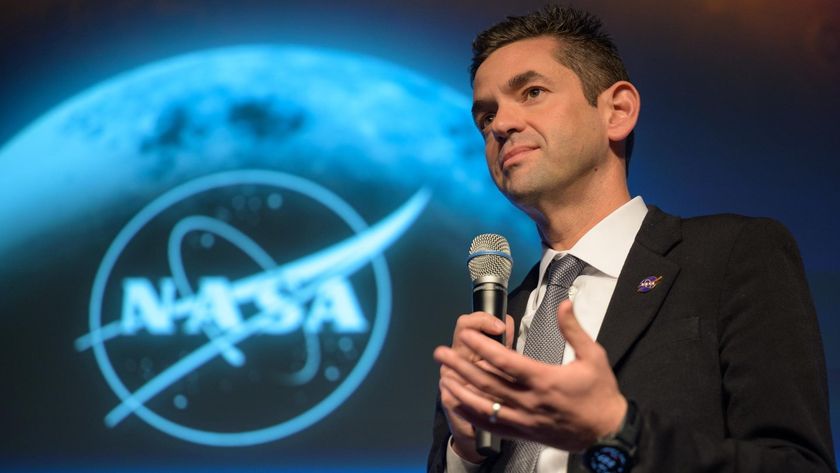 a man in a suit speaks into a microphone in front of a nasa emblem