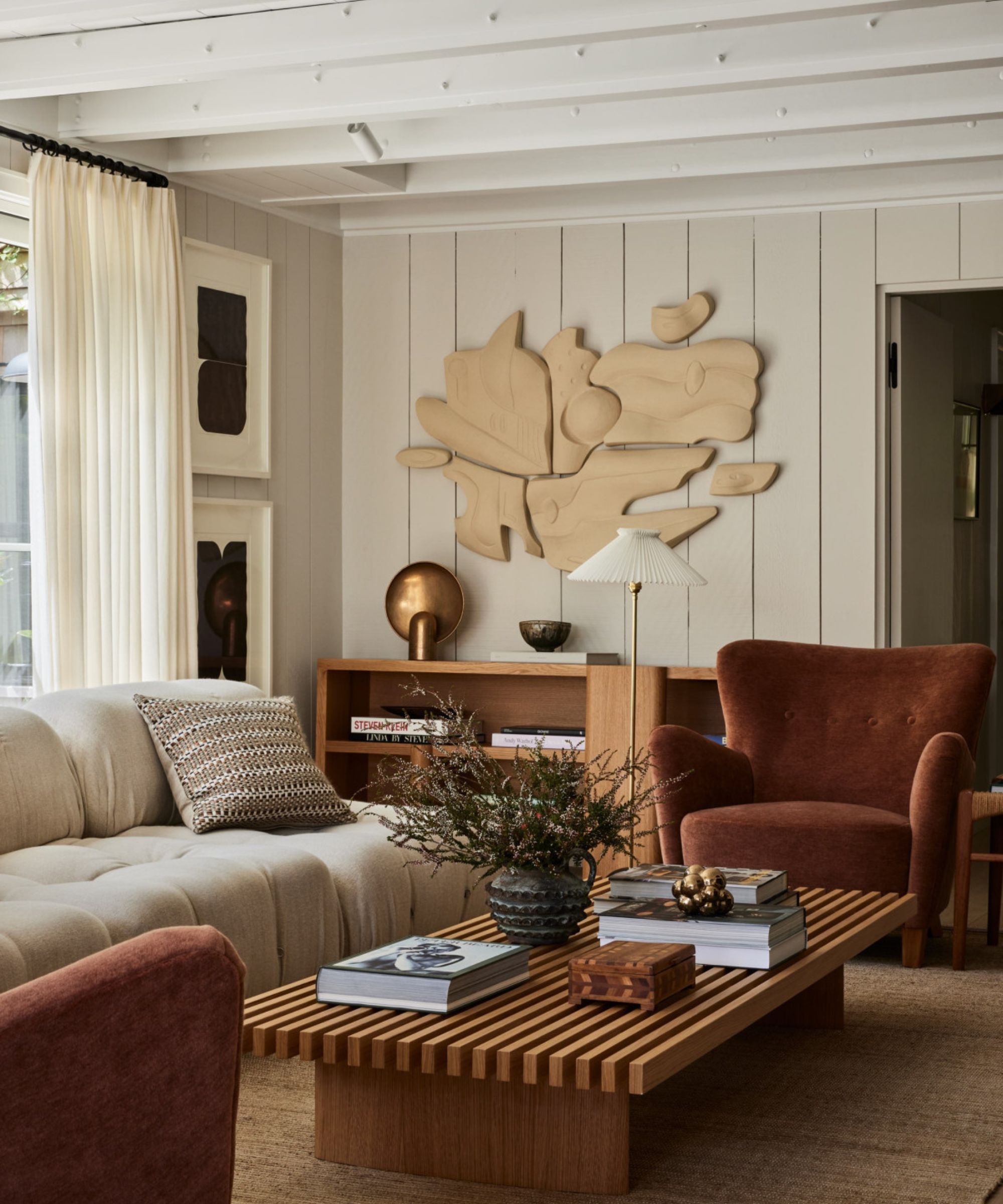 A bright, contemporary living space with cream wood-paneled walls, a slatted oak coffee table, and a sculptural tan wall installation above a terracotta armchair