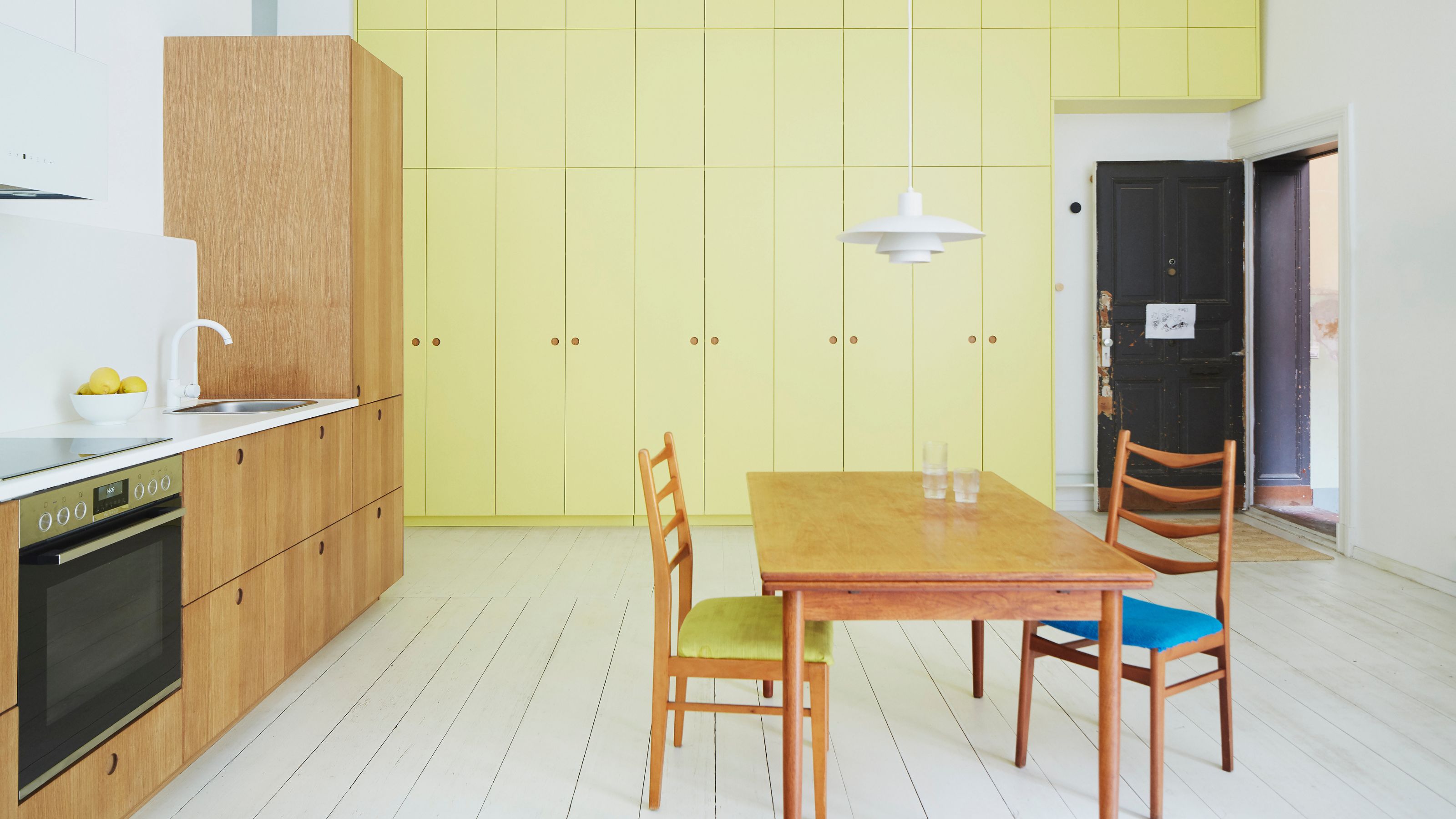yellow kitchen cabinets with timber kitchen cabinets, white painted floorboards, a timber table with two timber chairs, white countertops, white tap, and old door 