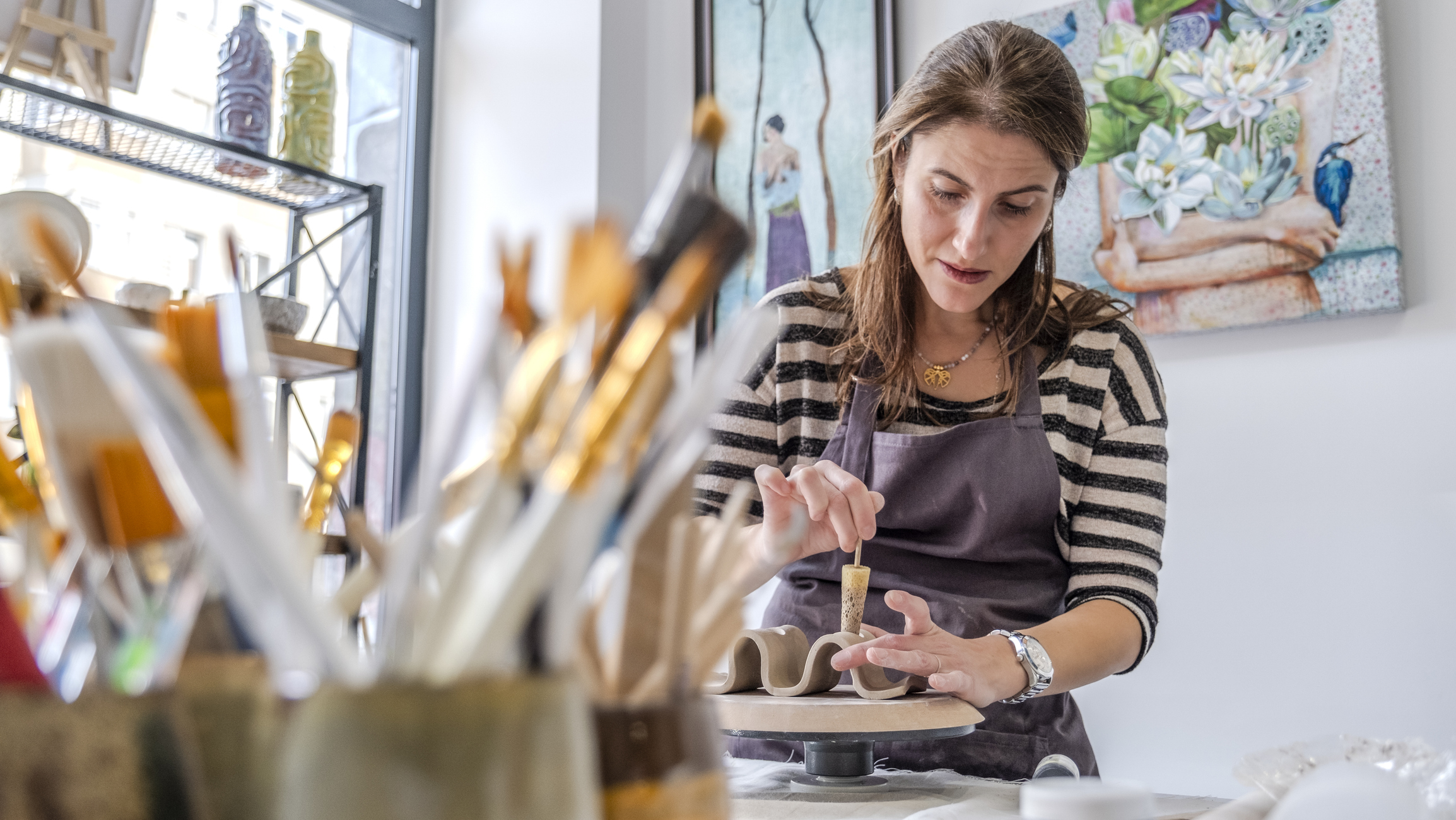 A shot of a middle-aged woman artist shaping clay with sculpting tools at a table in an art studio, with an environment adorned with artistic paintings in the background