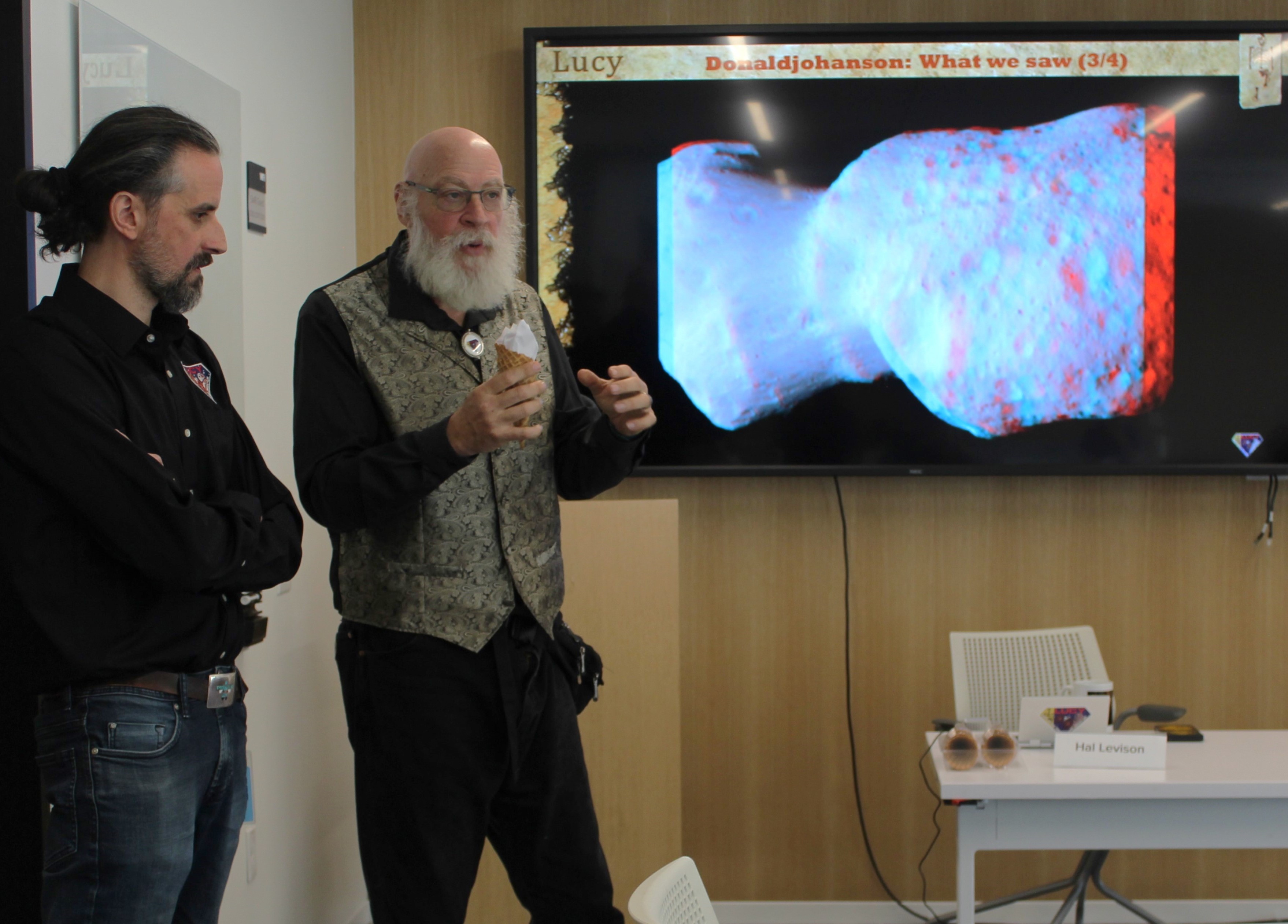 a bald man in a button-down shirt and bolo tie stands in front of a television screen with a photo of an irregularly-shaped rock on it