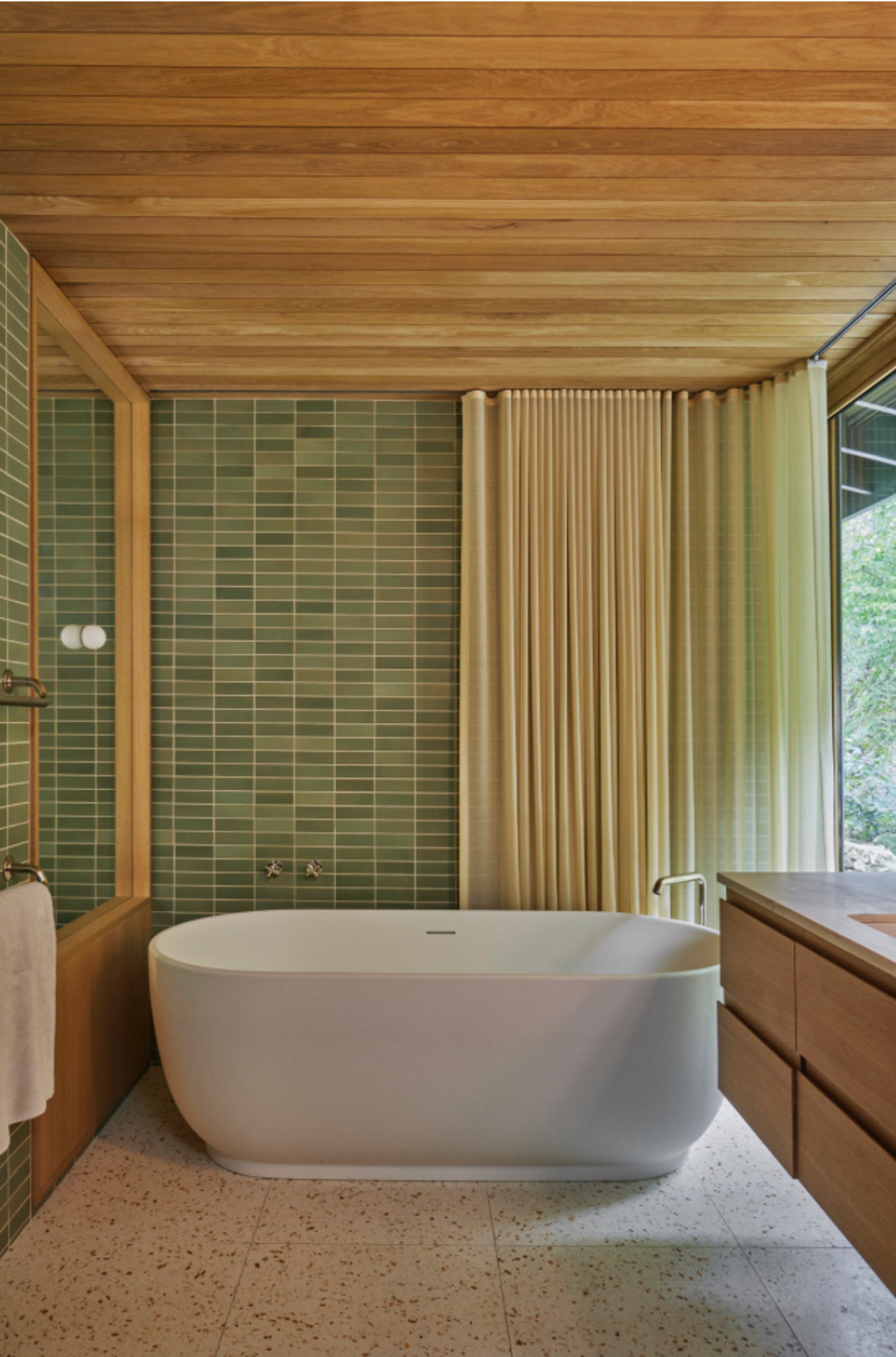 mid century modern bathroom with green wall tiles, timber clad ceiling, curtains, freestanding white bathtub, terrazzo floors, and floating timber vanity