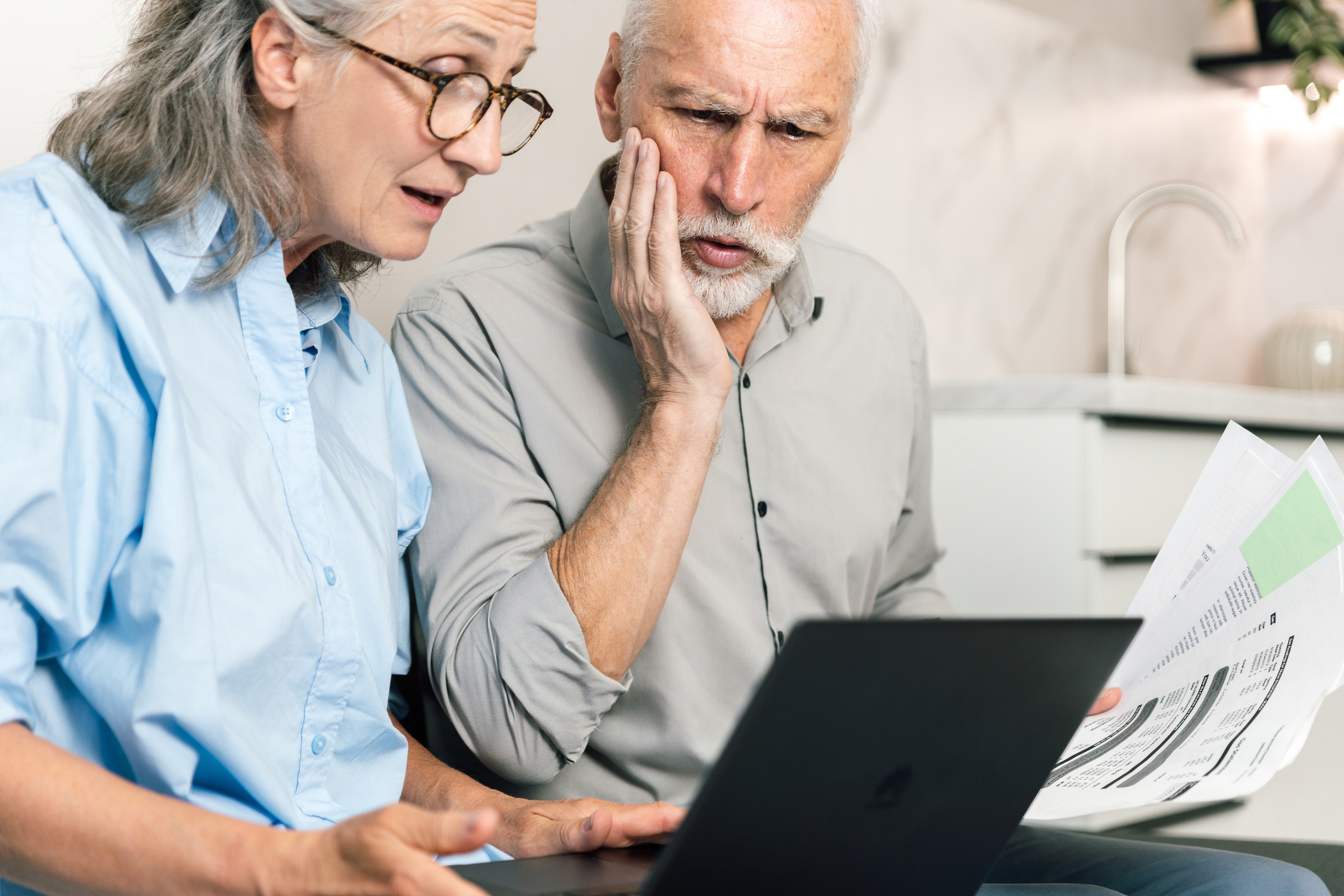 Sad and depressed senior couple discusses their finances while looking at a laptop. They check bank statements and utility bills, focusing on problems, managing their savings.