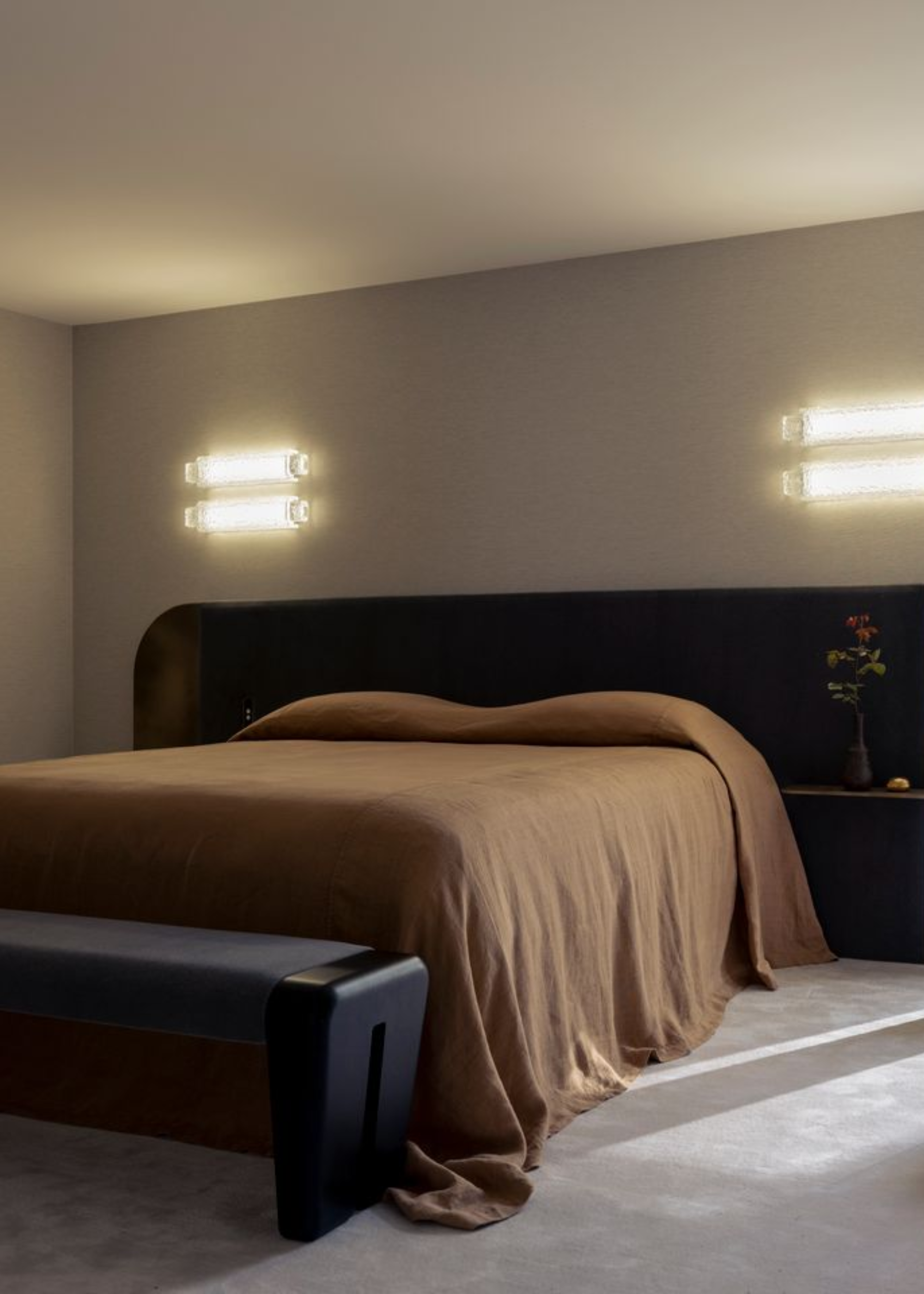 A dark beige bedroom with rectangular wall sconces, an oversized brown headboard with a floating side table, a wood bench, and a brown bedscape