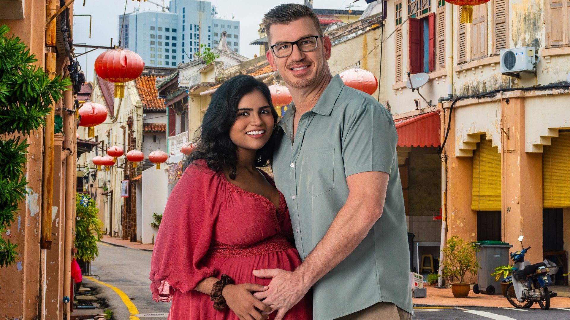 Jonathan and Ally in Malaysia. She's wearing a pink maternity dress and he's wearing blue short-sleeve button up. He has his hand on her pregnant stomach and they're both smiling.