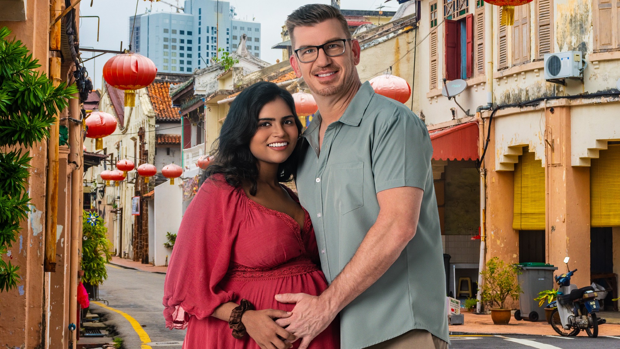 Jonathan and Ally in Malaysia. She's wearing a pink maternity dress and he's wearing blue short-sleeve button up. He has his hand on her pregnant stomach and they're both smiling.