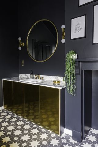 An example of dark bathroom ideas showing metallic basin unit doors, a gold mirror, gold wall lights and a marble worktop with patterned floor tiles