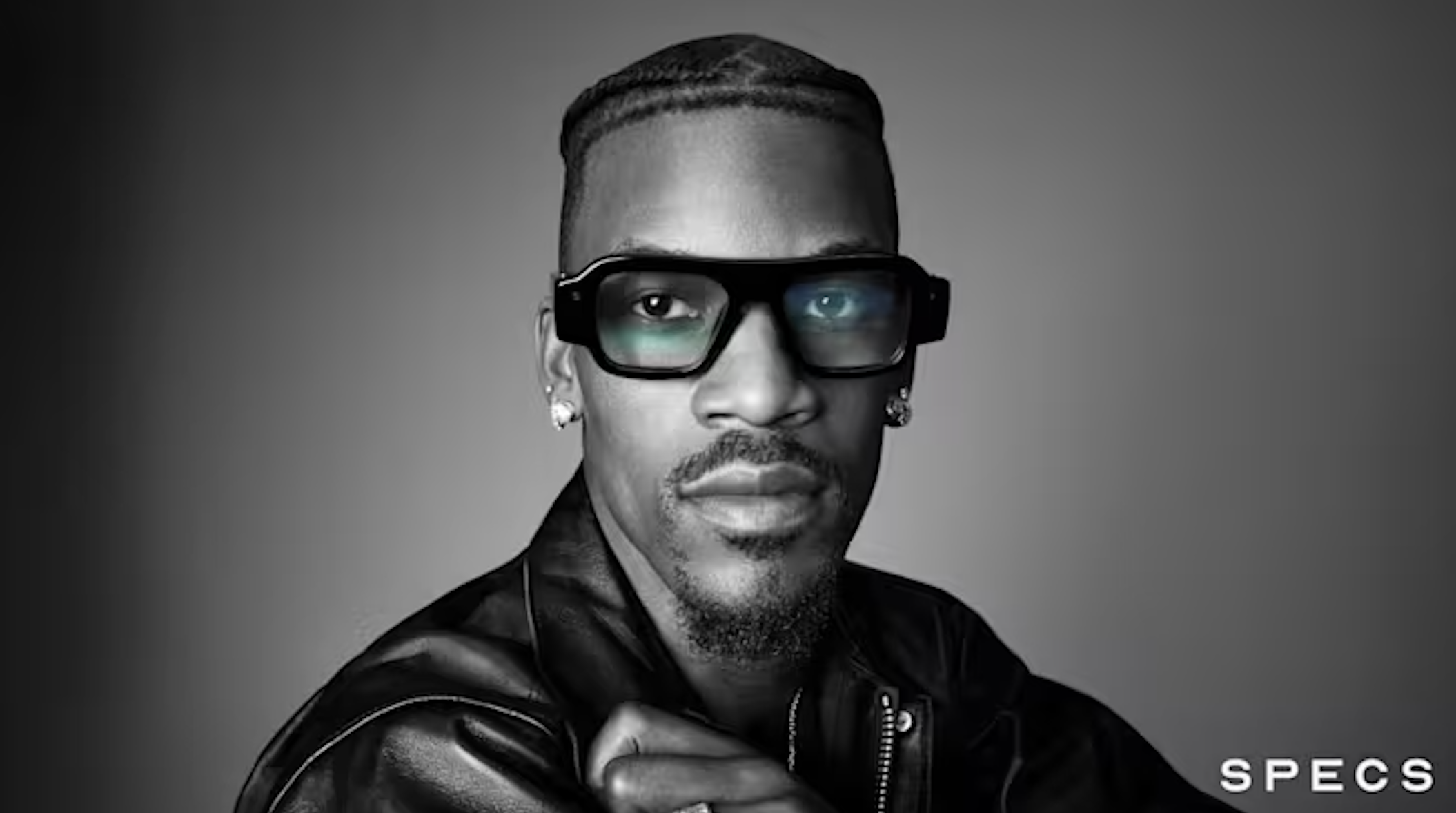 black-and-white, front-facing portrait of a man with braided hair, a goatee, and earrings, wearing a leather jacket and thick, black-framed glasses.