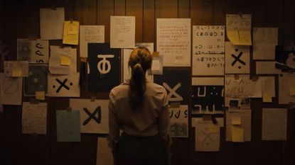 a woman with her back to the camera stands in front of a wall of papers and cryptic drawings