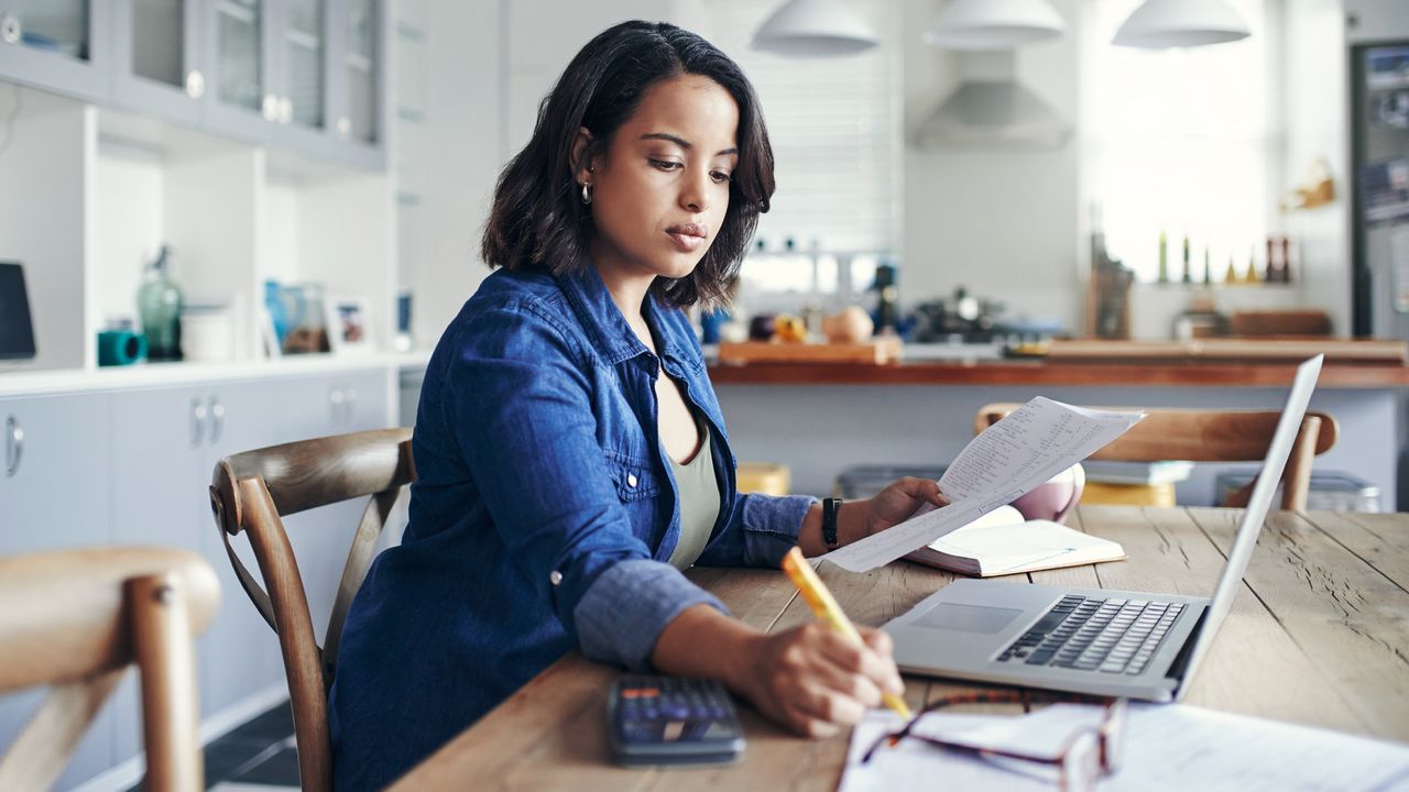 A woman works at her dining room table, making notes while her laptop sits open in front of her.