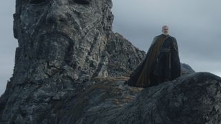 Still from season 1 of the Star Wars TV show Ahsoka. Baylan Skoll (human male, white hair and beard, wearing a long flowing brown cloak) stands tall as he searches for answers. He is standing on a mountain which has a face carved into it.