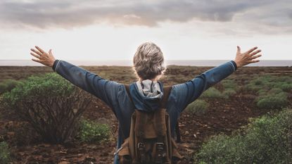 An older hiker holds her hands outstretched to the horizon.