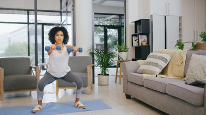 Woman in workout clothes doing a squat with two dumbbells in a stylish living room