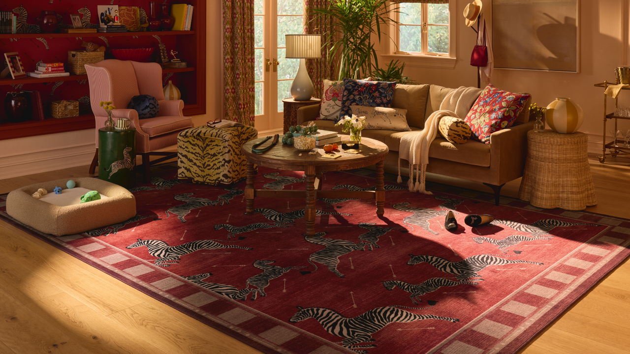 A maximalist living room with a bold red rug with jumping zebras on it, a couch with several pillows on top, a round coffee table, a printed ottoman, a dog bed, and a pink accent chair on the left side 