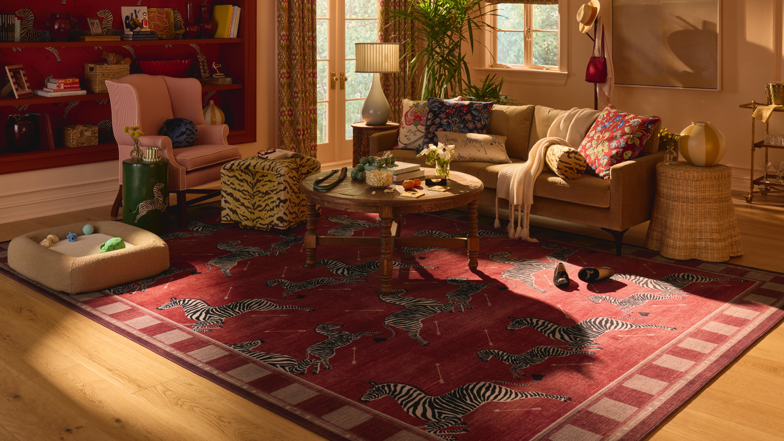 A maximalist living room with a bold red rug with jumping zebras on it, a couch with several pillows on top, a round coffee table, a printed ottoman, a dog bed, and a pink accent chair on the left side 