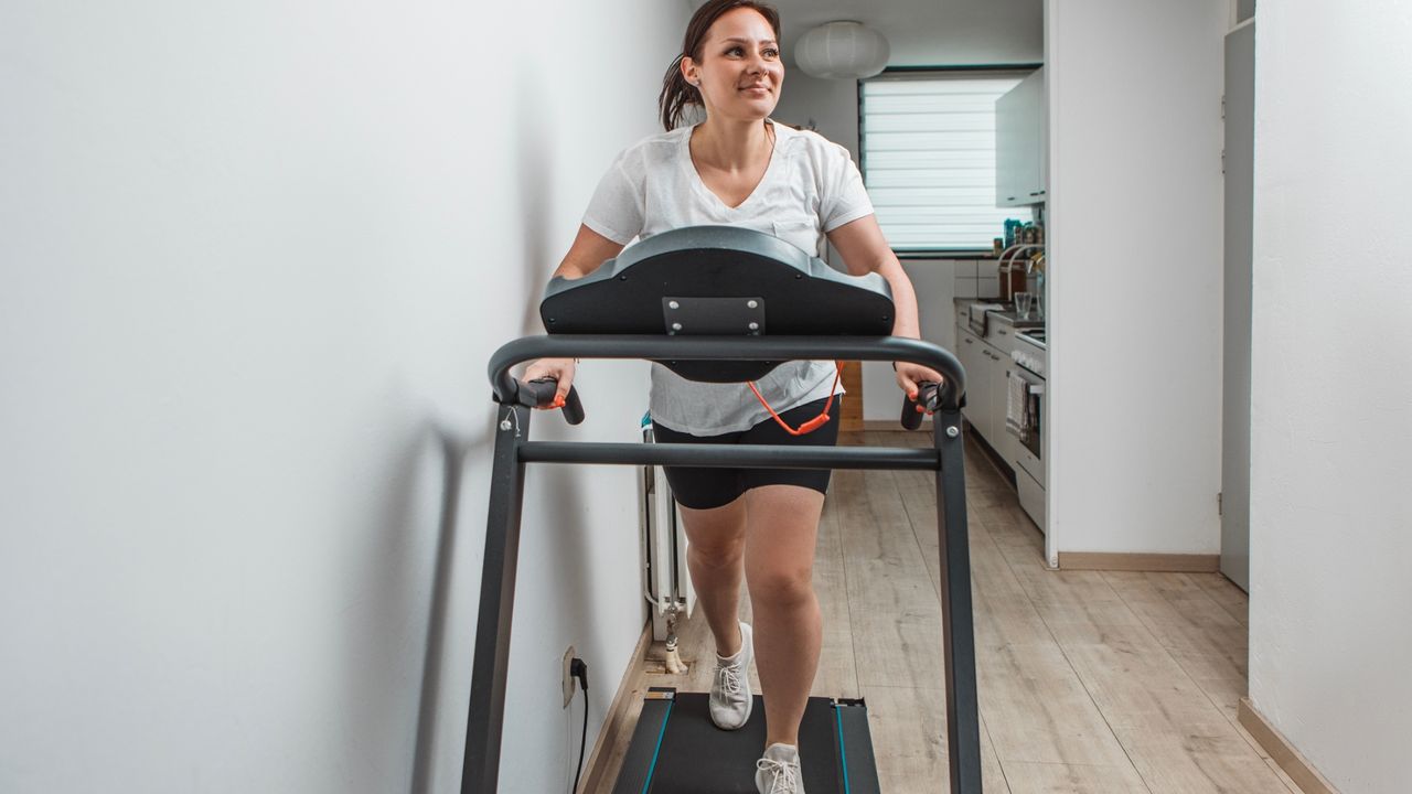 woman on a treadmill with a white walls on both sides, a wooden floor and a narrow kitchen behind her. she's wearing a white tshirt and black shorts and looking away from the camera smiling