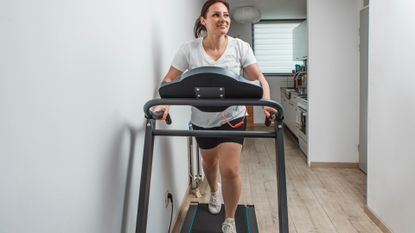 woman on a treadmill with a white walls on both sides, a wooden floor and a narrow kitchen behind her. she's wearing a white tshirt and black shorts and looking away from the camera smiling