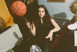 The Seattle Storm's Nika Mühl sitting in a desk chair wearing Under Armour sneakers while playing with a basketball.