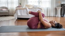 woman on a grey exercise mat lying on her back holding her knees to her chest. she's in a bedroom with a bed and windows behind her.