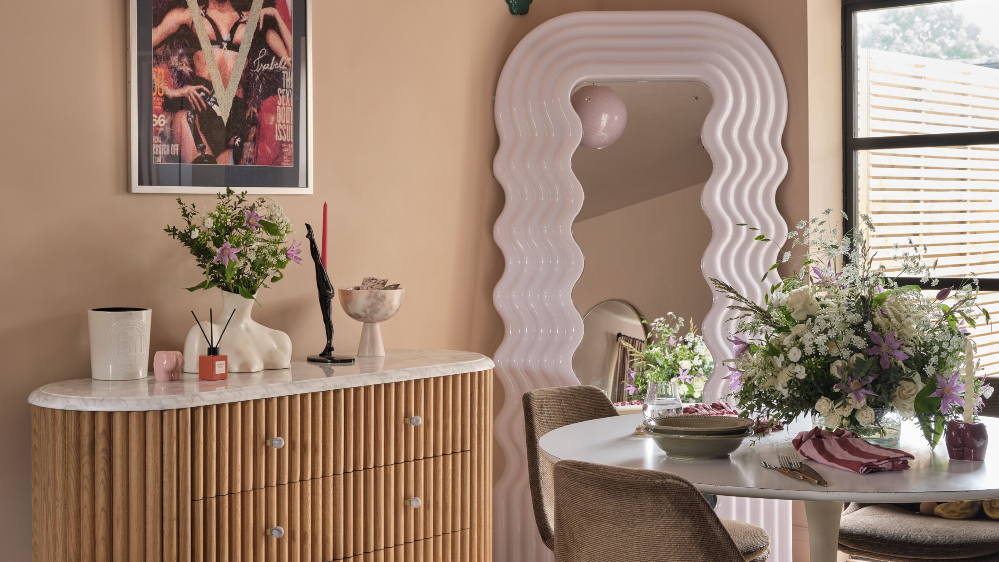 A dining room with a round table, a wavy full-length mirror and a reeded sideboard with a marble top