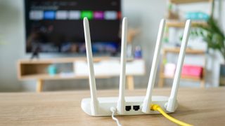 A wired, white-colored router sits on a wooden surface with its wireless antenna pointing upward. In the background is an out-of-focus smart TV displaying its home screen.