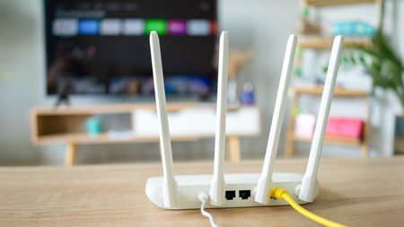 A wired, white-colored router sits on a wooden surface with its wireless antenna pointing upward. In the background is an out-of-focus smart TV displaying its home screen.