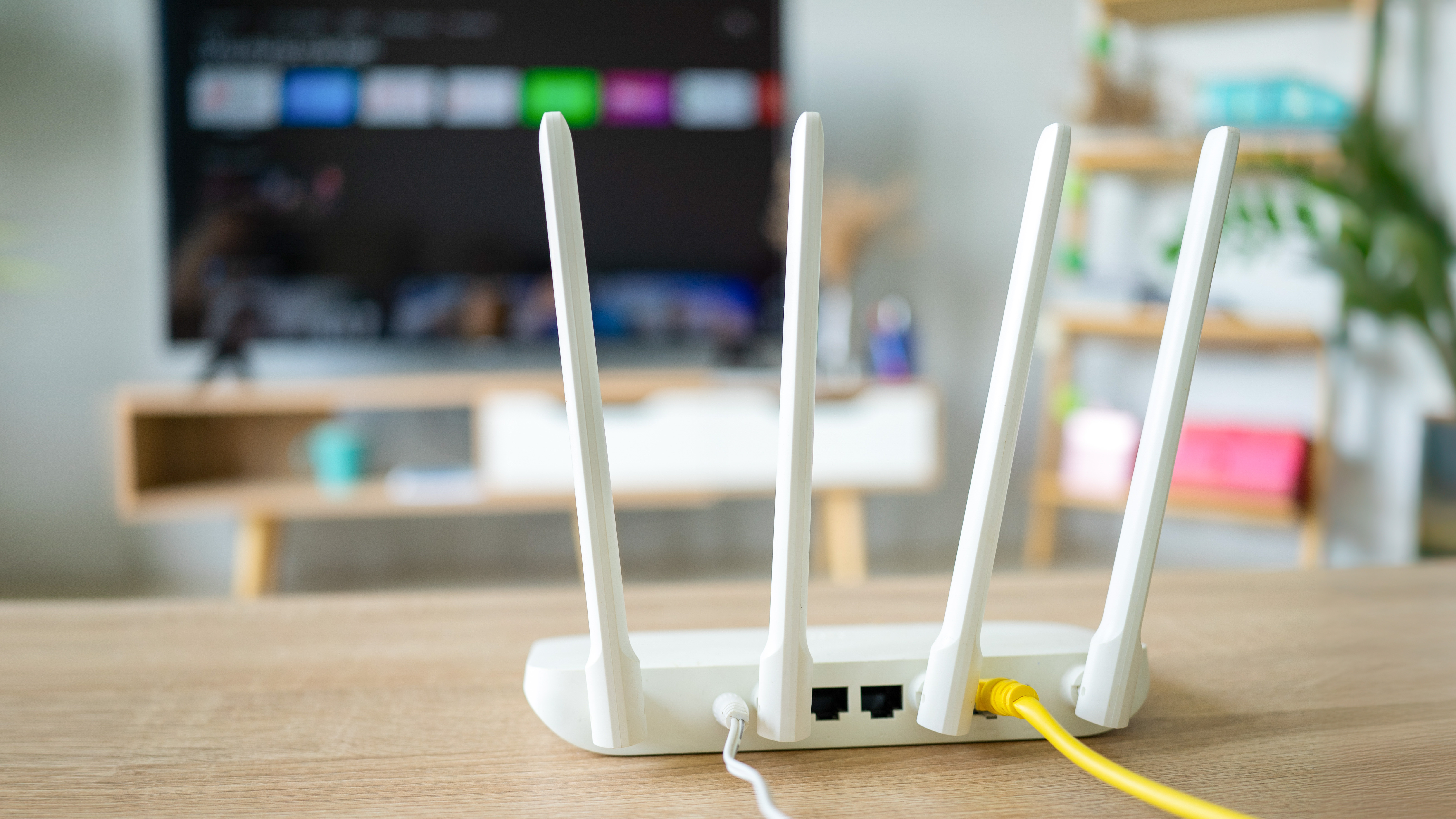 A wired, white-colored router sits on a wooden surface with its wireless antenna pointing upward. In the background is an out-of-focus smart TV displaying its home screen.