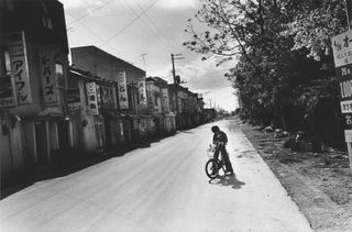 Black and white photograph of a young per-son riding a bicycle on a street.