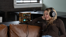 A woman with long blonde hair listening to Grado Signature S750 headphones on a brown leather sofa with a nice-looking amp on a unit behind the sofa.