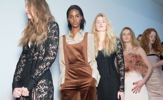 Models waiting in line for a fashion show to start, wearing a long, lace see-through dress and a retro-looking bronze jumpsuit, from the Topshop Unique A/W 2015 collection.