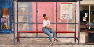 Dionne Brown as Queenie sitting at a bus stop.