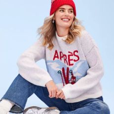 Blonde woman wearing red hat and 'Apres' snow jumper with blue jeans and background